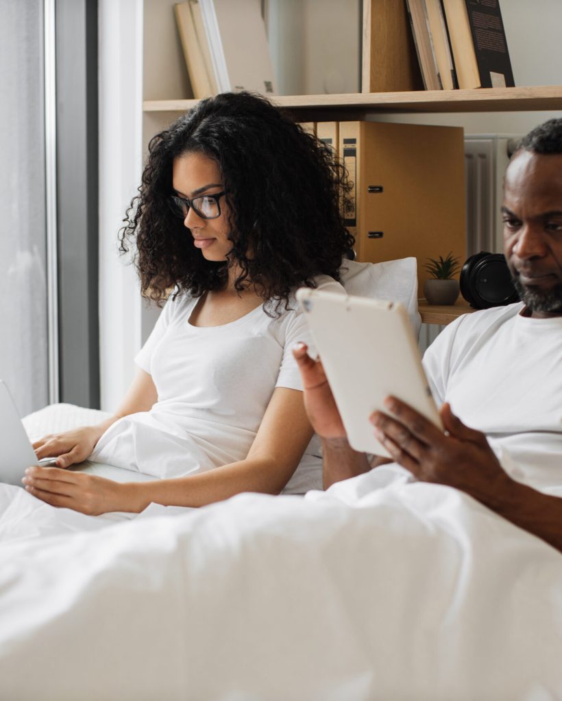 Pretty african american woman working on laptop in bed while focused man searching information via digital tablet. Young family finding work-life balance without negative impact on relationships.